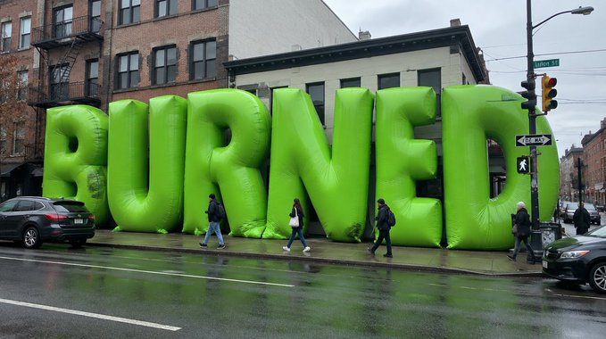 A giant inflatable green "PURVEY" sign occupies a city street corner, towering over pedestrians walking by in the rain, with the accompanying post referencing a cryptocurrency loss where 18.7763 ETH was converted into 8,743,672 $GRASS tokens that have since vanished, seemingly using the oversized "PURVEY" display as visual irony to mock the situation.