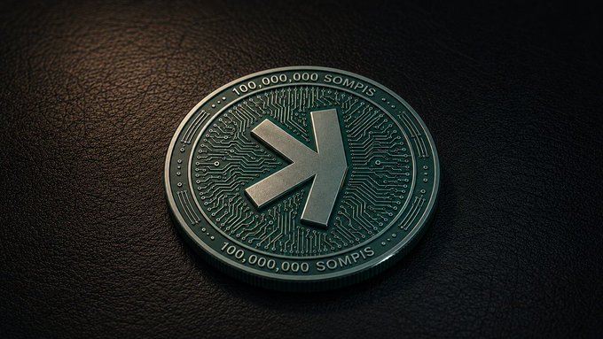 A close-up of a silver commemorative coin with a stylized 'K' logo in the center, inscribed with '100,000,000 SOMPIS' around the edges, resting on a dark textured surface.