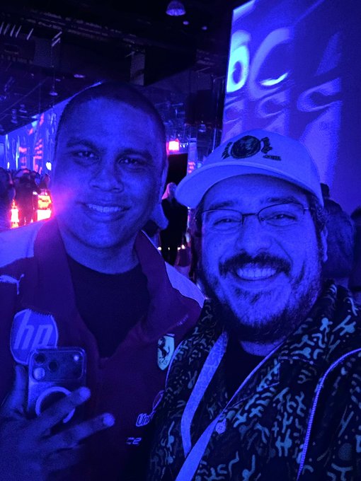 Two men smiling for a selfie in a dark, blue-lit environment; one wears a vest with an HP logo and holds a phone, while the other wears a white cap, glasses, and a patterned hoodie.