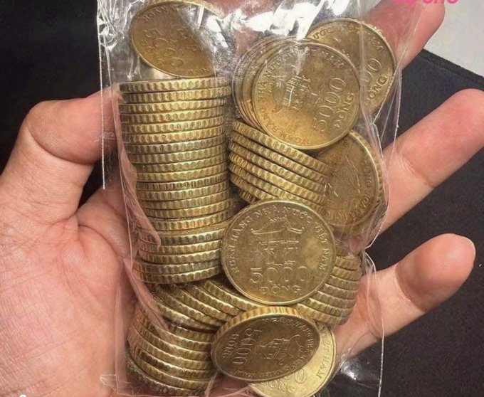 A hand holding a clear plastic bag filled with stacks of gold-colored coins marked "1000 DONG" and "5000".