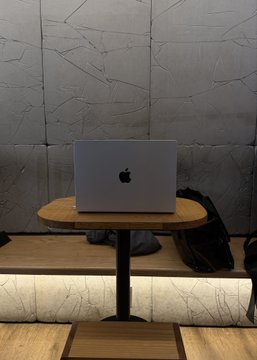 A silver Apple MacBook sits open on a round wooden bistro table in what appears to be a sleek, modern café or co-working space with stone-tiled walls and a matching bench. Beside the laptop, a black backpack rests on the bench, suggesting a remote work setup. The scene captures the relatable experience of working remotely—enjoying the perk of a quiet, lock-in-friendly space after noon, only to return to an overwhelming flood of Slack notifications waiting to be tackled.
