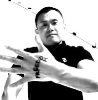 A black and white photo of a man wearing a black FILA shirt, holding his hands up toward the camera to display a tattoo on his knuckles.