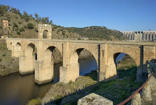A scenic photograph of a medieval stone arch bridge spanning a river, with rolling hills and a modern dam visible in the background.
