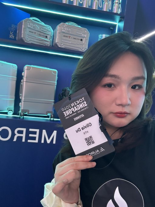 A young woman holds up a VIP pass reading "SILVERBACKER PERLESS","SILVER TIER - BLACK CARD HOLDER", and "Olivia Di XTH" with "KORUA" visible at the bottom, standing in front of a display of silver cases labeled "NEMO".