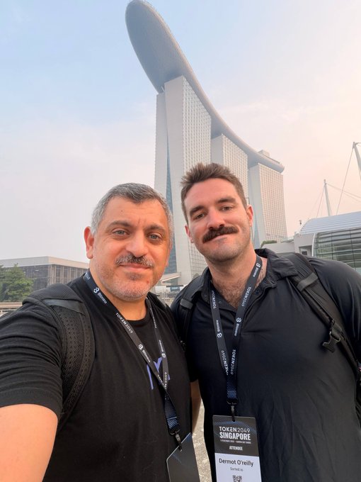 Two men taking a selfie in front of the Marina Bay Sands hotel in Singapore; one wears a lanyard labeled 'Dermot Greally' for the 'TOKEN 2049 SINGAPORE' event.
