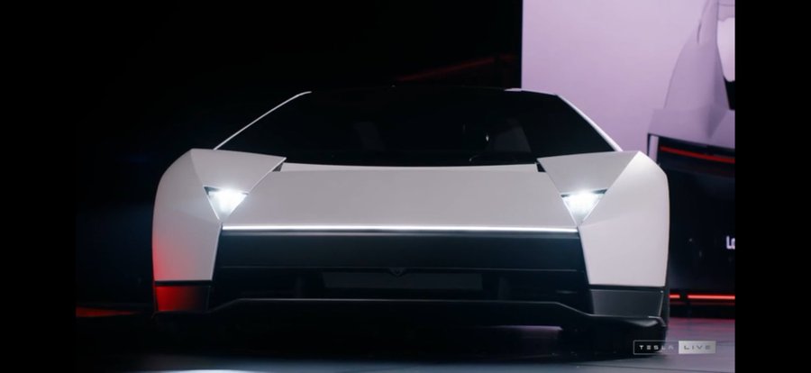A white Tesla concept vehicle displayed on stage with its headlights illuminated, featuring 'TESLA LIVE' branding in the corner.