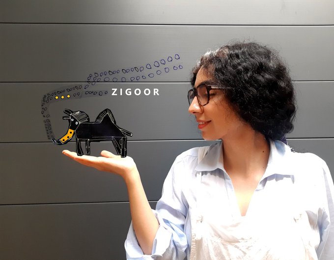 A woman with curly hair and glasses holds a small Zigoor figurine (a black creature with a yellow horn) in her palm against a gray wall displaying the 'ZIGOOR' logo.