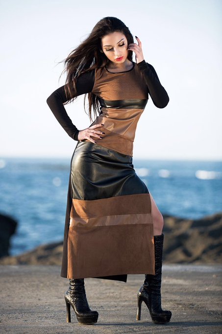 A woman poses on a rocky shore by the ocean, wearing a brown and black paneled dress with tall black platform boots.