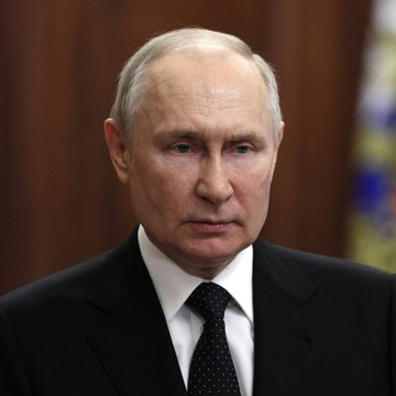 A portrait photograph of Vladimir Putin wearing a dark suit and tie, with a blurred Russian flag visible in the background.