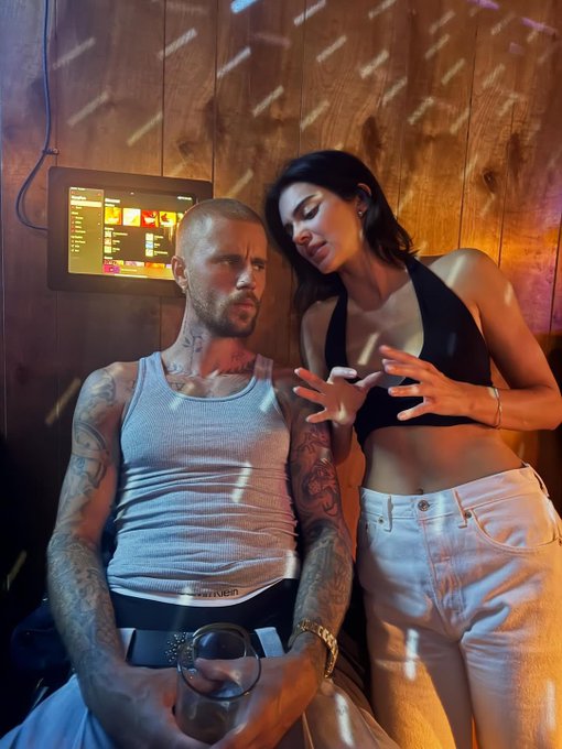 A man in a white tank top and a woman in a black halter top and light skirt stand closely together in a dimly lit room with a screen in the background.