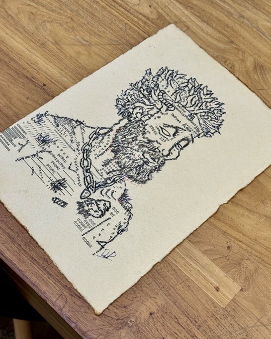 A hand-drawn portrait of a bearded figure with a crown of leaves on textured paper resting on a wooden table, signed by the artist.