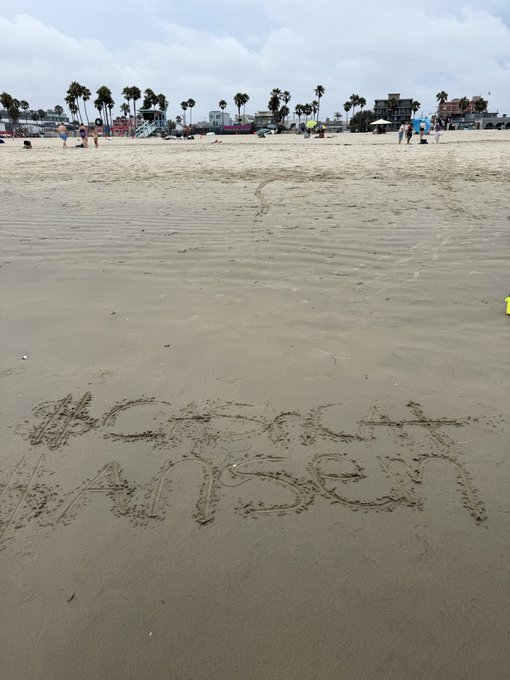 Someone has written "#cashcat got no ankles" in the sand at Venice Beach, where the photographer is working to earn free bags for crypto tokens $CASHCAT and $ANSEM on a community "family day" event. The image captures the beach setting with palm trees and distant crowds, illustrating the casual, meme-driven promotional atmosphere tied to the cryptocurrency project.