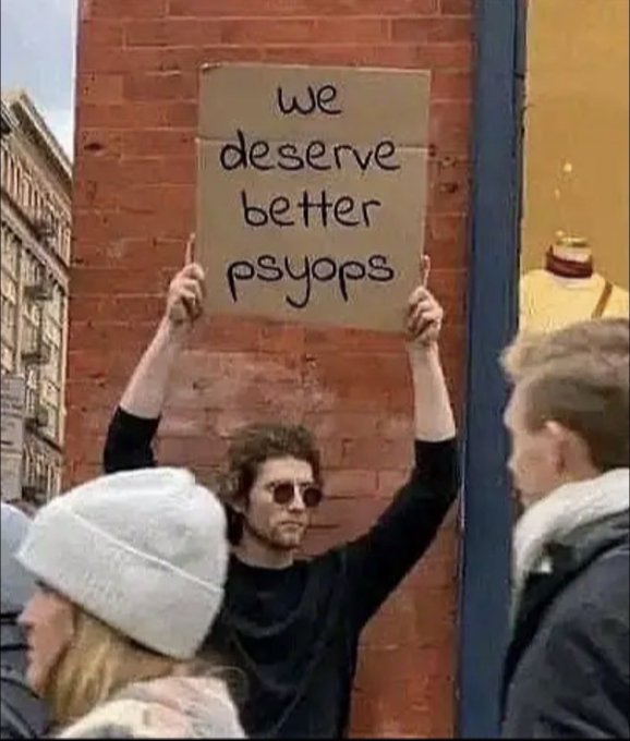 A man at a protest holds up a cardboard sign reading 'we deserve better psyops' against a brick wall.