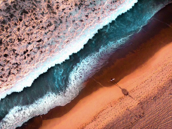 An aerial photograph of a sandy beach with turquoise waves crashing onto the shore, showing a single person's shadow cast on the sand near the water's edge.