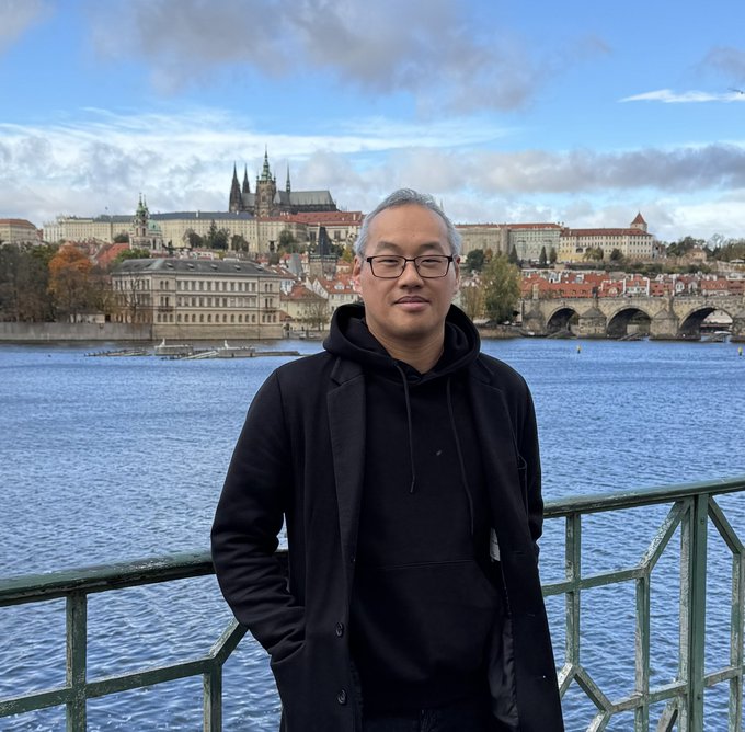 A man in a black coat and hoodie stands before a scenic view of Prague Castle and the Charles Bridge spanning the Vltava River. The caption "Your collector" suggests a personal, perhaps sentimental, connection to the location, framing the photo as a keepsake from a meaningful trip.