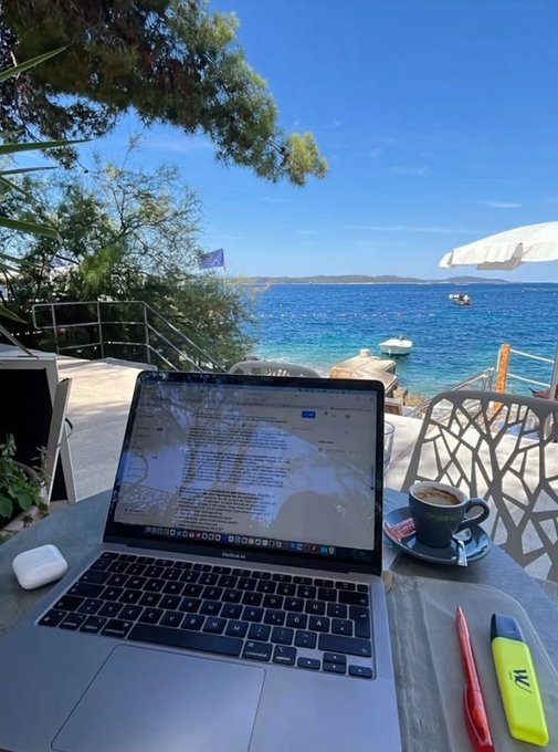 A MacBook laptop sits on an outdoor table overlooking a bright blue sea and rocky coastline, accompanied by a coffee cup and yellow/red highlighters.