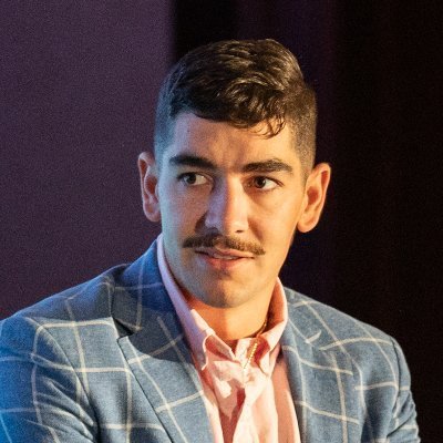 A portrait photograph of a young man with dark hair and a mustache, wearing a blue plaid blazer over a pink shirt, looking off to the side.