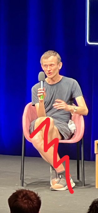 Vitalik Buterin sits cross-legged on a pink chair holding a microphone on stage, with a red squiggly line drawn over his legs to humorously represent Ethereum's price chart plunging from $4,900 to $1,700.