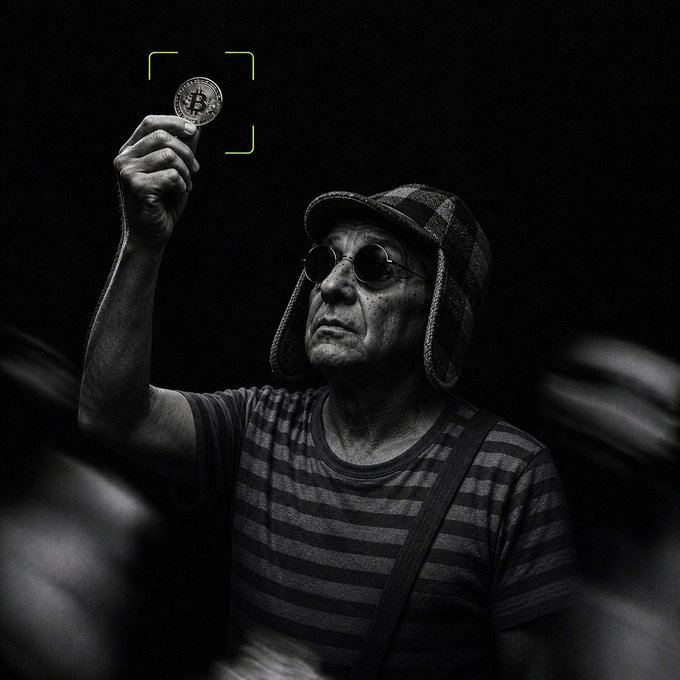 Black and white photo of an older man wearing a striped shirt, sunglasses, and a trapper hat, holding up a Bitcoin coin with a yellow focus frame graphic around it.