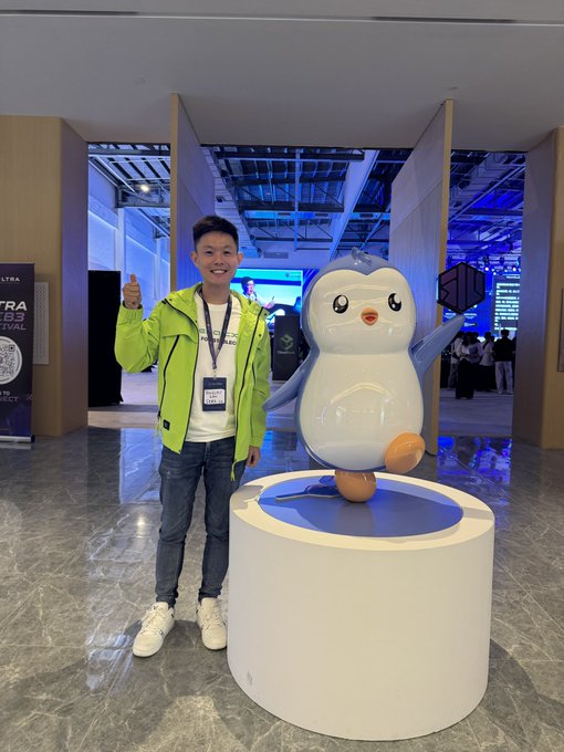 A young man in a green jacket giving a thumbs up next to a large penguin mascot statue on a white pedestal at what appears to be a convention or trade show.