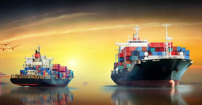 Two large cargo ships loaded with shipping containers are sailing on calm water during a vibrant sunset, with a small airplane visible in the sky on the left.