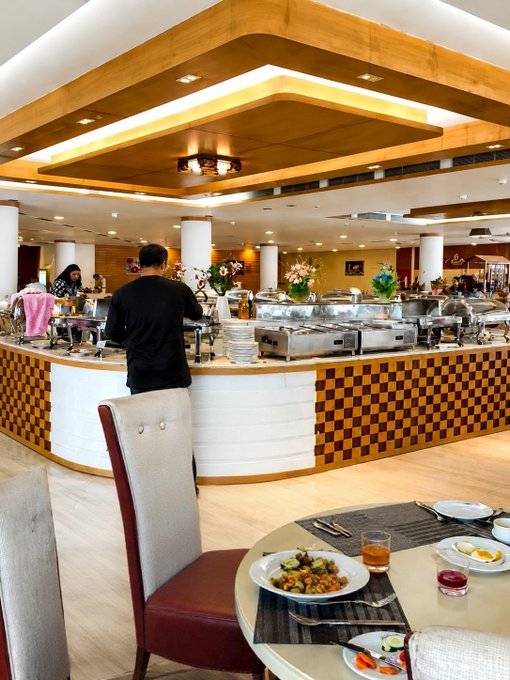 An interior view of a hotel or restaurant buffet featuring a curved serving counter with checkered orange and white tiles, chafing dishes, and a few patrons. A dining table in the foreground holds plates of food including vegetables and eggs.