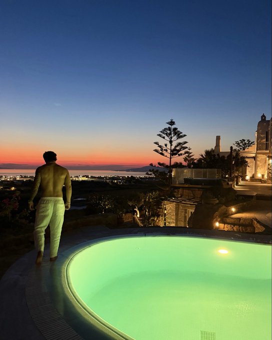 A shirtless man in white pants stands at the edge of an illuminated infinity pool overlooking a Mediterranean coastline at sunset, embodying the luxurious, picturesque lifestyle implied by "Euro maxxing."