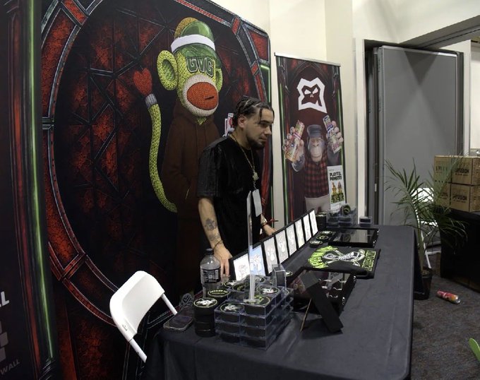 A man works behind a merchandise table at a convention booth decorated with a large mural of a sock monkey wearing a green cap marked “SWS”; shirts, hats, patches, and displays are spread across the table.