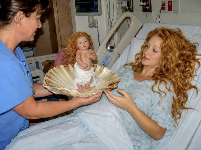 A nurse presents a small red-haired doll wrapped in a white towel inside a large seashell to a smiling woman with long red hair in a hospital bed.