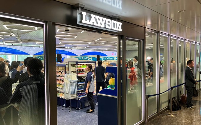 The image shows the storefront of a Lawson convenience store in Tokyo, with customers browsing inside and a businessperson waiting in line outside, corresponding to the announcement that an early August proof-of-concept test for JPYC stablecoin payments will take place at a single Lawson store in Tokyo to verify POS-integrated transactions.