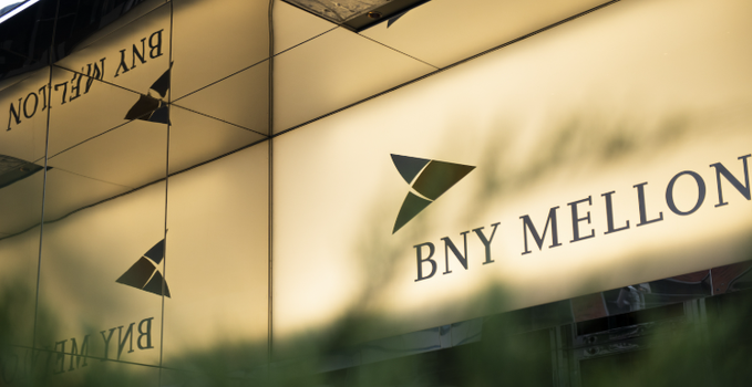 The image displays a gold-illuminated sign featuring the "BNY MELLON" logo and name on the exterior of a building, with reflections of the signage visible in adjacent glass panels. This visual reinforces the article's announcement of the historic custodian bank entering the cryptocurrency staking sector through its partnership with Galaxy Digital.