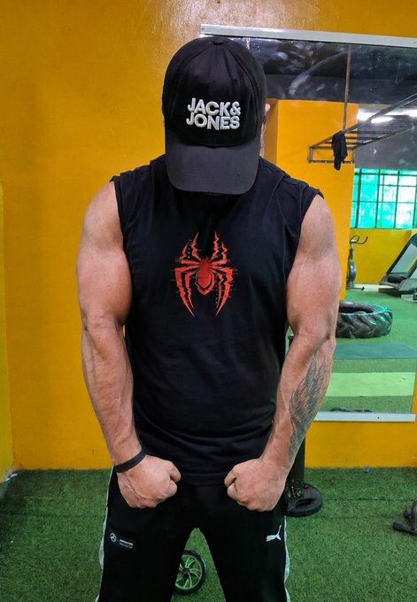A muscular man in a gym wearing a black 'Jack & Jones' cap, a black sleeveless shirt with a red spider logo, and black Puma pants, posing with his head down.