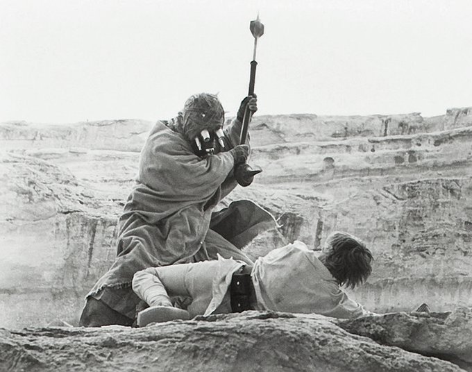 A black and white photograph of a Tusken Raider from Star Wars raising a weapon (a gaffi stick) over a fallen figure in a desert landscape.
