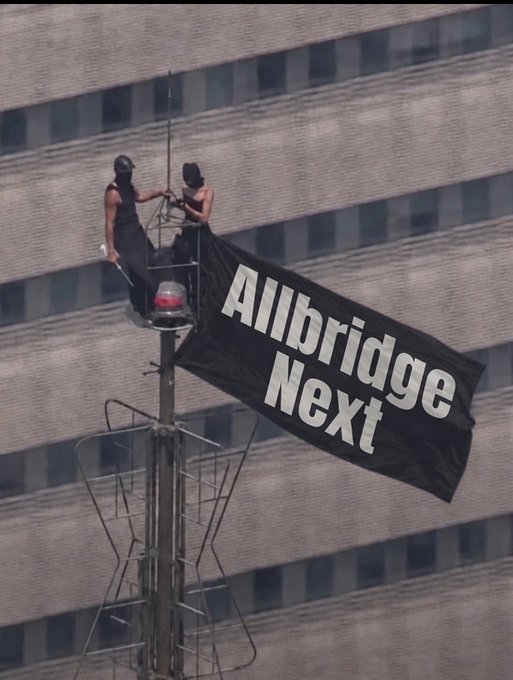 Two people in black clothing and caps standing on a pole-mounted platform attached to a building, displaying a large black banner reading 'Allbridge Next'.