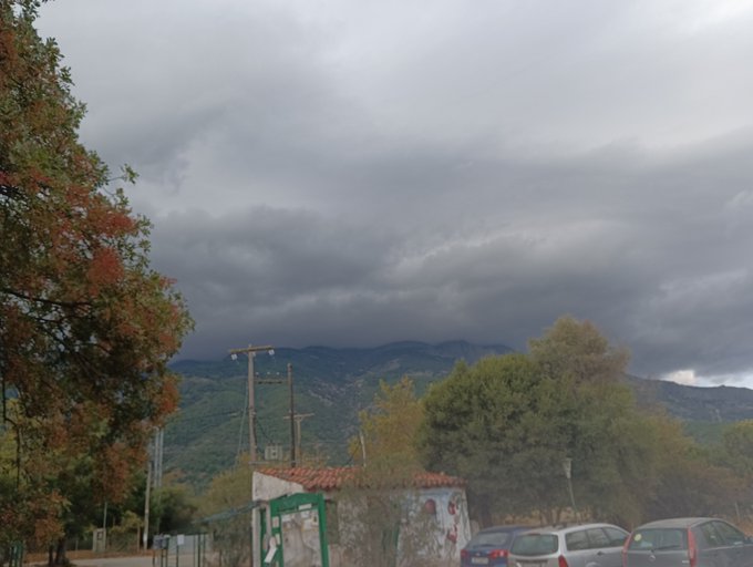 An outdoor scene showing a cloudy, overcast sky above mountains and trees, with a small building, utility poles, and parked cars in the foreground.