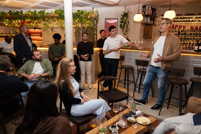 A man addresses a seated group inside a warmly lit bar or restaurant, with a “HAPPY HOUR” sign visible behind the counter.