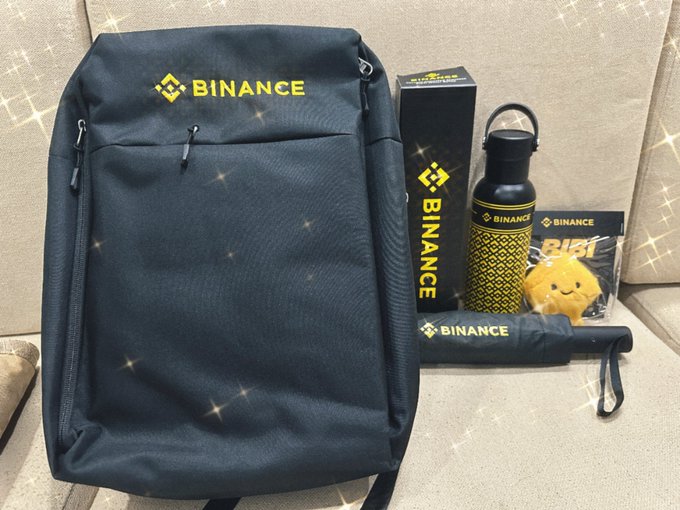 A collection of Binance-branded merchandise including a backpack, water bottle, box, umbrella, and small yellow mascot toy arranged on a beige couch.