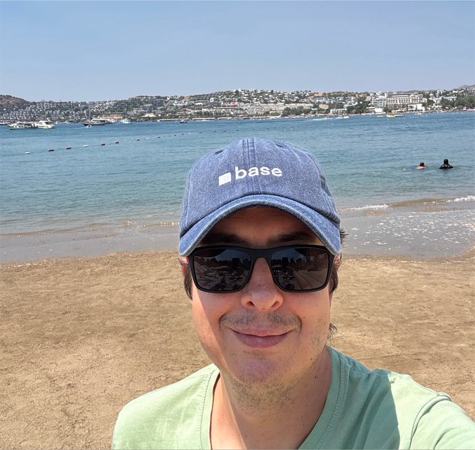 A man takes a selfie on a sandy beach, wearing a blue cap with the word 'base' printed on it, black sunglasses, and a light green shirt, with ocean and coastal buildings visible behind him.