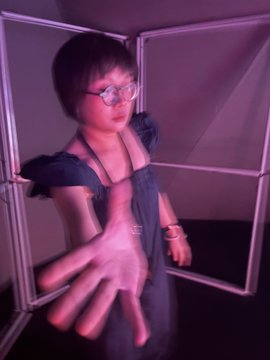 A young person with glasses wearing a denim top, extending their hand toward the camera in a pink-tinted indoor setting.