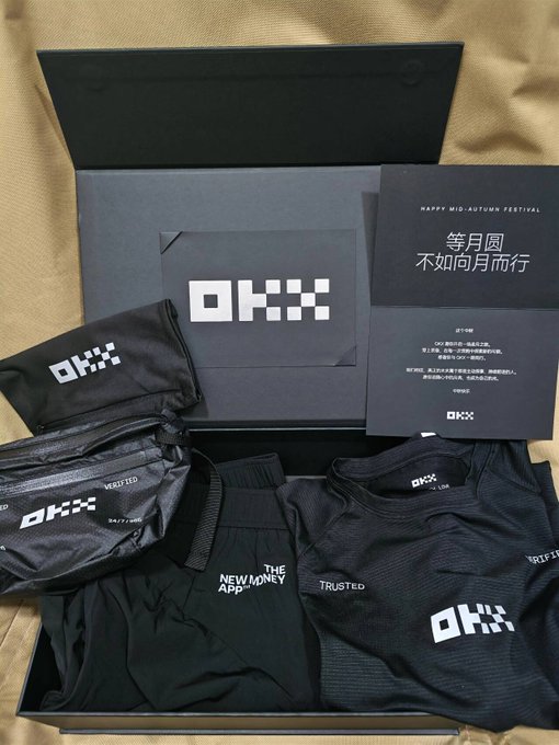 An open OKX branded gift box containing black athletic apparel, a drawstring bag, and a card reading 'HAPPY MID-AUTUMN FESTIVAL' with Chinese characters '等月圆 不如向月而行' (Wait for the full moon, better to walk toward it).