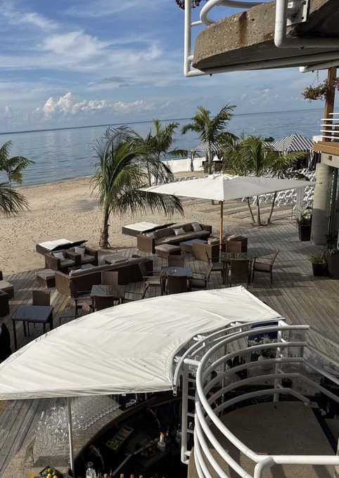 A view from a beachside bar/restaurant showing a wooden deck with white umbrellas, tables, and chairs, overlooking a sandy beach with palm trees and calm blue ocean waters.