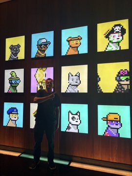 A person stands taking a photo of a wall display featuring illuminated CryptoPunks-style pixel art characters arranged in a colorful grid.