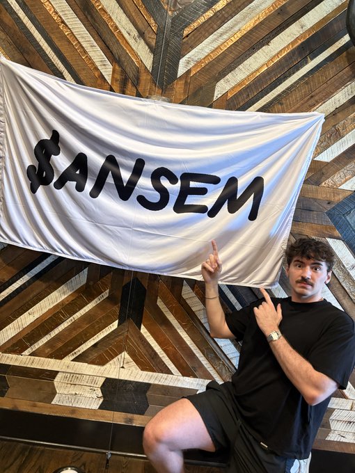 A man in a black outfit points upward at a white banner reading "$ANSEM" against a wooden chevron-patterned wall.