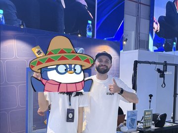 A bearded man in a white t-shirt gives a thumbs-up while standing next to a penguin mascot character wearing a sombrero and a red scarf.