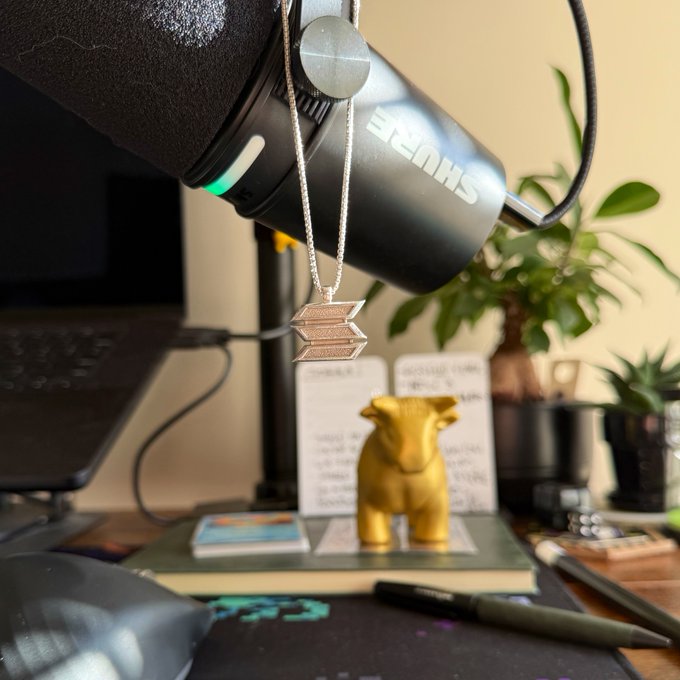 A photograph of a Shure microphone with a silver chain pendant hanging from it, positioned above a small yellow bull figurine and handwritten notes on a cluttered desk with plants in the background.