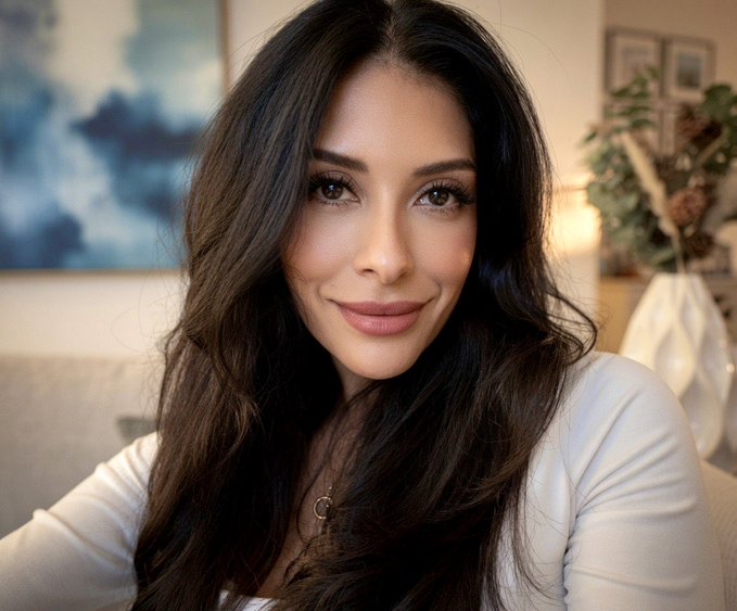A woman with long dark wavy hair smiles softly at the camera, wearing a white top and a small pendant necklace, with a blue painting and plant visible in the background.