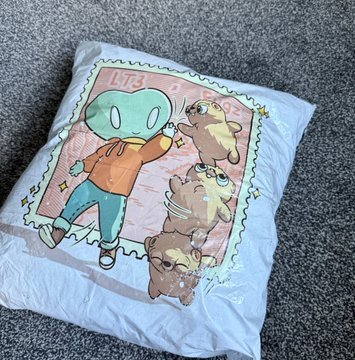A white cushion or fabric piece featuring cartoon-style illustrations of an alien-like character and three animals (a capybara, cat, and dog) with the text 'CUTE ALIENS' displayed on a stamp-like design.