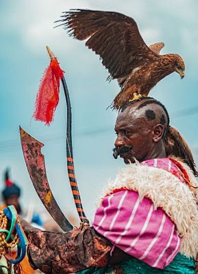 A portrait of a traditional falconer in a pink garment with a sheepskin shoulder piece, holding a large curved blade in one hand while a large bird of prey perches on his head, set against a blue sky.