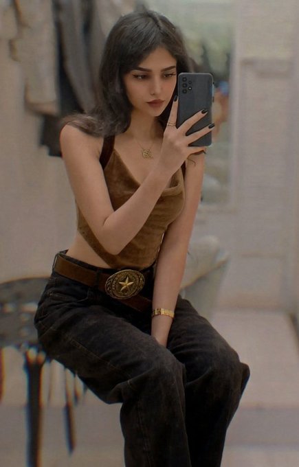 A young woman taking a mirror selfie while wearing a tan top, dark jeans with a star belt buckle, and jewelry.