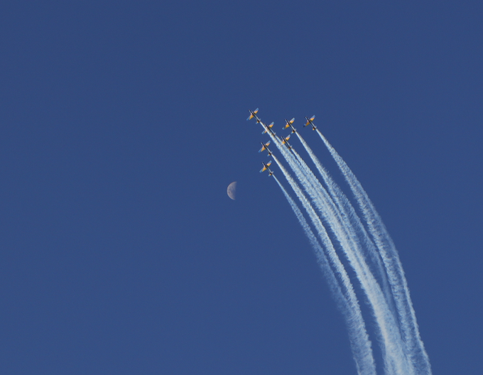 Aerial photograph of six aircraft flying in formation leaving white contrails across a deep blue sky, with the crescent moon visible in the distance.